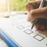 A hand holding a pen marks a checkbox on a clipboard with a checklist, ensuring every detail is attended to. A window with a blurred view of greenery is in the background.