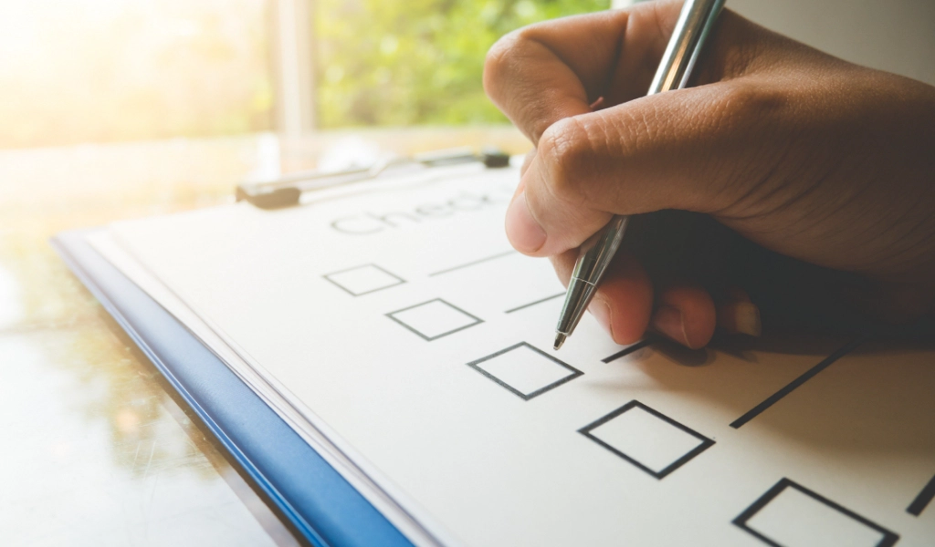 A hand holding a pen marks a checkbox on a clipboard with a checklist, ensuring every detail is attended to. A window with a blurred view of greenery is in the background.