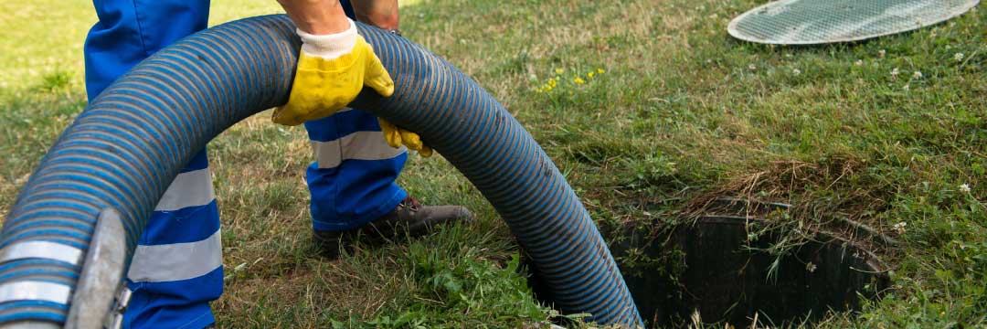A worker from the local septic tank service holds a hose while standing near an open septic tank on a grassy area.