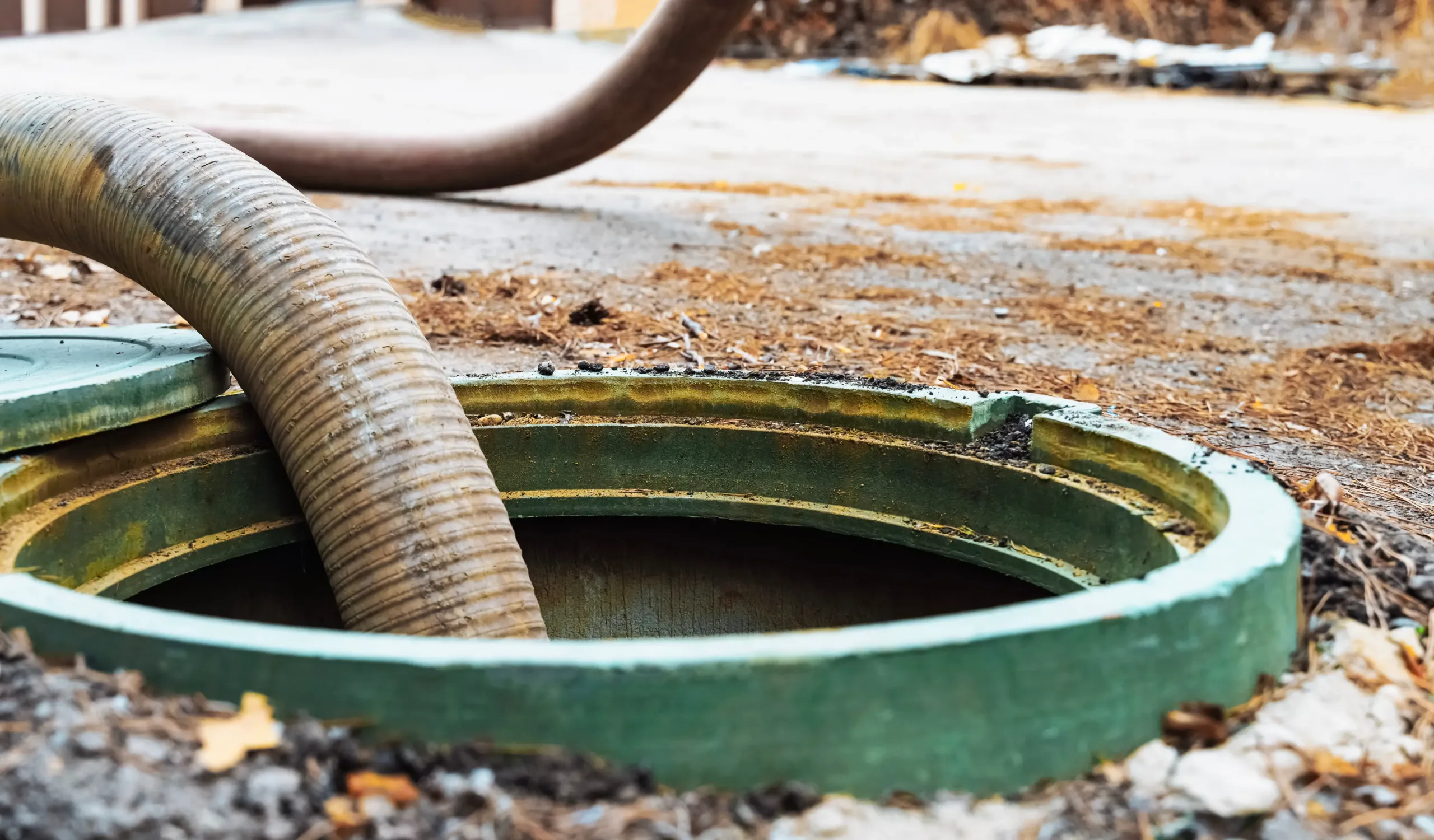 Close-up of a septic tank being serviced with a large vacuum hose inserted into the open green lid, surrounded by dirt and debris on the ground.